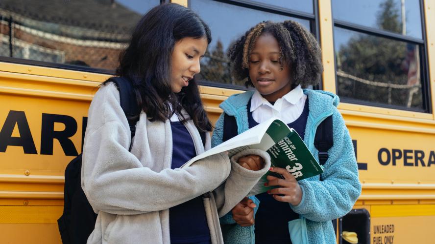 Students look at a textbook while standing near a school bus.